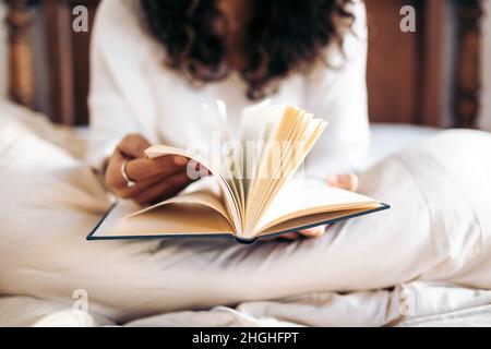 Nicht erkennbare junge Frau, die die Seiten eines Buches dreht, während sie im Bett liest, das mit der Bettdecke bedeckt ist Stockfoto