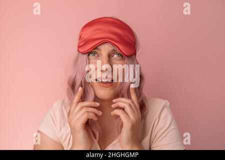 Positive Frau lächelt zieht rote Seide Schlafmaske erwacht in guter Stimmung nach dem Sehen angenehme Träume in der Nacht genießt Morgenzeit gegen Rosa Stockfoto