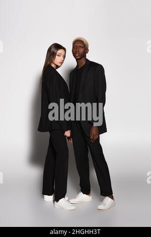 In voller Länge Ansicht von trendigen interracial paar in schwarzen Anzügen und weißen Sneakers auf grau mit Schatten Stockfoto