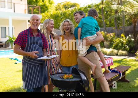 Porträt einer fröhlichen kaukasischen Familie aus drei Generationen, die im Garten gemeinsam grillen Stockfoto