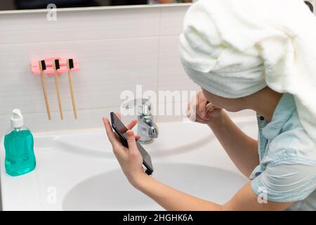 Eine junge hübsche Frau mit einem Handtuch auf dem Kopf in einem hellen Badezimmer mit einer Zahnbürste und einem Smartphone in der Hand bürstet ihre Zähne und kommuniziert mich Stockfoto