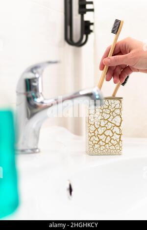 Eine Frauenhand steckt eine hölzerne Zahnbürste in ein Glas mit einem goldenen Muster im Badezimmer vor dem Hintergrund eines Wasserhahns und glänzender Fliesen. Selektiv Stockfoto