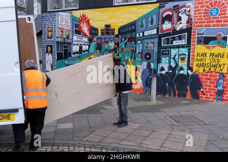 Zwei Männer tragen Vorräte aus einem weißen Lieferwagen und kommen an einem Wandgemälde vorbei, das an die Unruhen in Brixton im Jahr 1981 und den systematischen Rassismus und die Schikanen der Polizei gegen die schwarze Gemeinde Brixton am 21st. Januar 2022 in der Railton Road, London, England, erinnert. Das Kunstwerk der Künstler Jacob V Joyce, Monique Jackson, Ailsa Yexley, Buki Bayode und Sola Olulode wurde durch Crowdfunding finanziert und zeigt den 40th. Jahrestag der Aufstände in Brixton 1981, als die Unruhen vom 10th., 11th. Und 12th. April 1981 auf der Brixton Railton Road begannen, bekannt als die Front Line .. (Vollständige Beschriftung in „zusätzliche Informationen“). Stockfoto