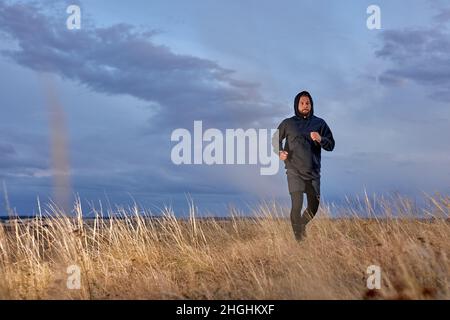 Mann joggt im Feld in schwarzer Sportkleidung, üben Training an der frischen Luft am Morgen. Bärtiger kaukasischer Athlet Kerl ist motiviert, konzentriert Stockfoto