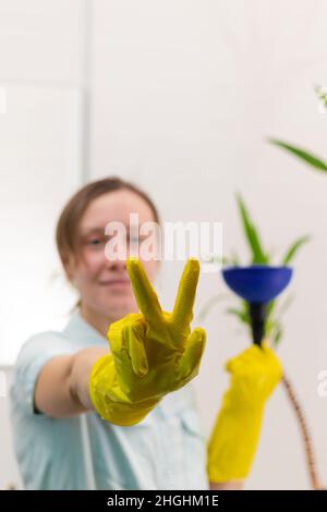 Eine junge Putzfrau in gelben hygienischen Gummihandschuhen in einem hellen Badezimmer mit einem Stößel, um die Verstopfung in ihren Händen zu beseitigen. Selektiver Fokus. Portra Stockfoto