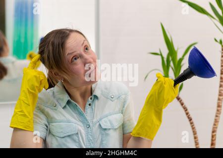 Eine junge Putzfrau in gelben hygienischen Gummihandschuhen in einem hellen Badezimmer mit einem Stößel, um die Verstopfung in ihren Händen zu beseitigen. Selektiver Fokus. Portra Stockfoto