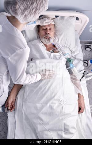 Draufsicht auf älteren Mann, der auf dem Bett liegt und an Krankheit leidet, weibliche Krankenschwester, die die Temperatur überprüft und die Hand auf die Stirn legt Stockfoto