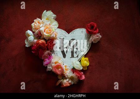 Herzförmige Holztafel mit LIEBE darauf geschrieben, umrandet mit Blumen auf einem dunkelroten Hintergrund. Stockfoto