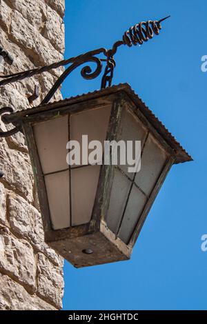 Alte, verzierte Laterne aus Eisen hängt vor einer rauhen Steinmauer vor dem starken blauen Himmel Stockfoto