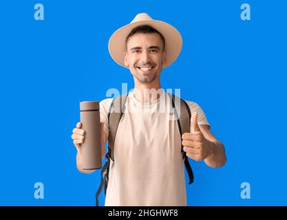 Männlicher Tourist mit Thermoskanne, die auf farbigem Hintergrund Daumen hoch zeigt Stockfoto