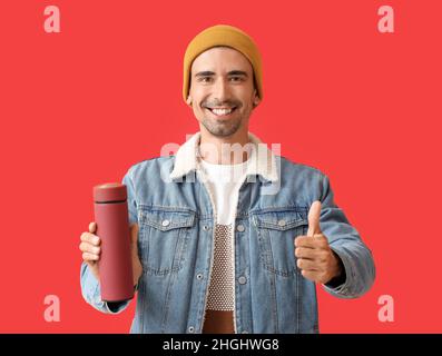 Hübscher Mann mit Thermoskanne, der auf farbigem Hintergrund Daumen hoch zeigt Stockfoto
