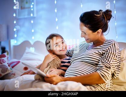 Du hast eine gute ausgewählt. Eine kurze Aufnahme einer attraktiven jungen Schwangeren, die ihre Tochter auf einem Tablet mit einer Geschichte zur Schlafenszeit vorliest. Stockfoto