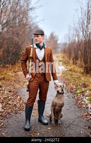 Schöner grauer Jagdhund mit Besitzer während eines Jagdtages. Herbstsaison, Jäger. Im Freien. Junge kaukasische starke Männchen in trendigen braunen Anzug zu Fuß Stockfoto