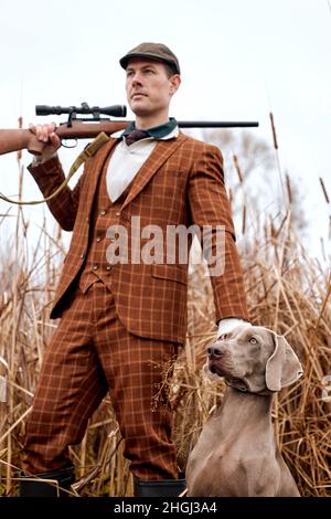 Aufmerksamer, starker, netter Jagdhund Weimaraner, der darauf wartet, vom gutaussehenden kaukasischen Wildhüter-Besitzer-Mann im trendigen, stylischen braunen Anzug das Kommando zu bekommen, zu Fuß Stockfoto