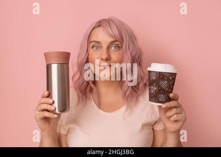 Porträt einer positiven kaukasischen Frau, die wiederverwendbare Thermobecher mit Einweg-Papierkaffeebecher vergleicht. Zero Waste und umweltfreundliches Konzept Stockfoto