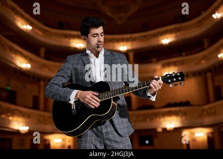 Junge Erwachsene Musiker spielen akustische Gitarre, Musik-Konzept, spielen ein Instrument. Schöner talentierter Kerl in grauen klassischen Anzug Musik. Stockfoto