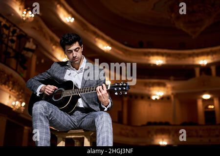 Talentierter Mann sitzen auf dem Stuhl spielen Gitarre auf der Bühne, auf Leistung. Schöner Kerl in formalen klassischen eleganten grauen Anzug Musik. Kunst, Menschen, mu Stockfoto