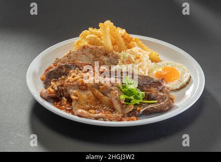 Traditionelle gemischte Grillplatte Gegrilltes Lammkotelett, Hähnchenschnitzel, Hühnerhacken, pommes frites, Eier und Salat in weißem Gericht auf dunkelgrauem Hintergrund Stockfoto