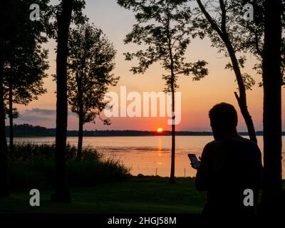 Silhouette einer Person, die ein Mobiltelefon benutzt, mit schönem Sonnenuntergang über dem See. Stockfoto