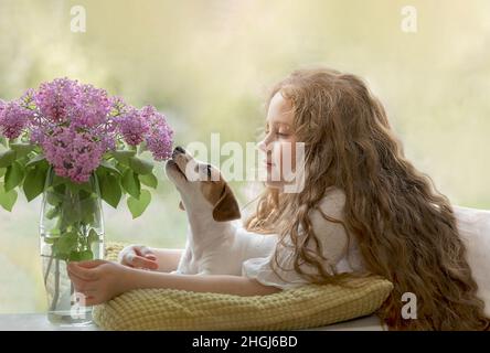 Lockiges Mädchen umarmt den Hund am Fenster. Freundschaft, Fürsorge, Liebe und glückliches Kindheitskonzept. Stockfoto