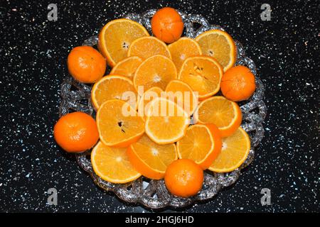 Nahaufnahme von leuchtend orangefarbenen Scheiben und ganzen Orangen, Satsuma-Früchten auf einem hübschen Glas-Servierteller auf einem schwarzen Küchentisch Stockfoto