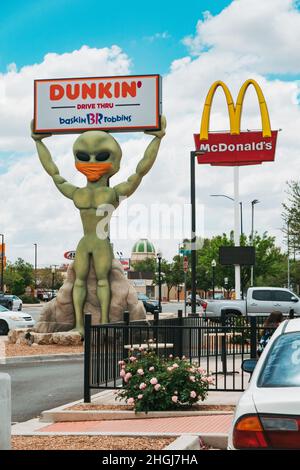 Eine Alien-Statue hält ein Dunkin' Donuts-Schild in Roswell, New Mexico, während sie eine Gesichtsbedeckung trägt. McDonalds UFO Restaurant ist sichtbar hinter Stockfoto