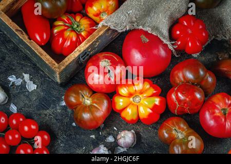 Draufsicht auf dunkle Tischfläche mit bunten hausgemachten Bio-Tomaten, Knoblauch, Holzkiste. Hausgemachtes Gemüse Erntekonzept Stockfoto