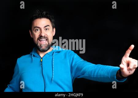 Junger Erwachsener mit Bart und spitzen Haaren in einem blauen Sweatshirt, der mit offenem Mund direkt in die Kamera blickt und Freude und Glück ausdrückt Stockfoto