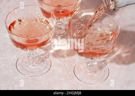 Rosé-Sekt oder Champagner, der aus der Flasche in Kristallgläser auf rosa Marmorhintergrund gießt. Sommerdrink-Konzept. Minimale kreative Komposition mit Kopierplatz. Stockfoto