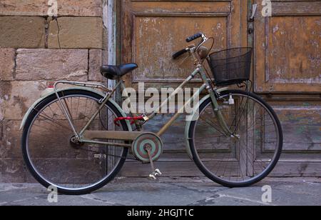 Ein klassisches, altes Vintage-Fahrrad mit Korbkorb liegt an einer verwitterten Wand und Tür in Lucca, Italien Stockfoto