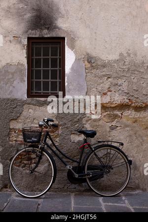 Ein altes Fahrrad mit Metallkorb ruht in Lucca, Italien, gegen eine verwitterte Zementwand Stockfoto