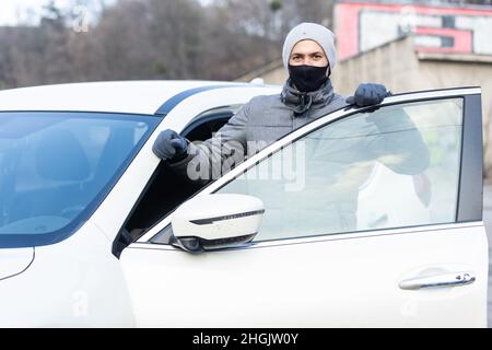 Junge Erwachsene Brünette Mann in chirurgische medizinische Maske, Jacke posiert auf der Straße der Stadt in der Nähe des Autos. Außenaufnahme. Stockfoto