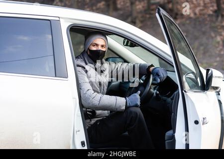 Junge Erwachsene Brünette Mann in chirurgische medizinische Maske, Jacke posiert auf der Straße der Stadt in der Nähe des Autos. Außenaufnahme. Stockfoto