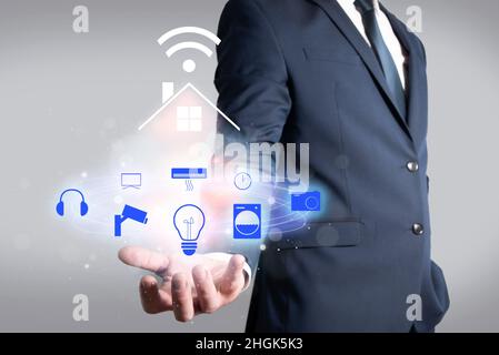 Mann in der Jacke hält ein Haus, das über WLAN mit verschiedenen elektronischen Geräten verbunden ist. Heimautomation im Haus Stockfoto