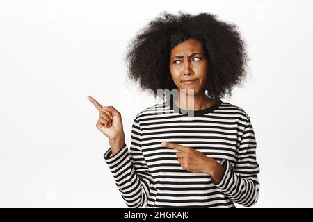 Bild einer jungen afroamerikanischen Frau, die zweifelhaft aussagt, zeigt und starrt, links mit enttäuschtem, skeptischem Stirnrunzeln, weißem Hintergrund Stockfoto