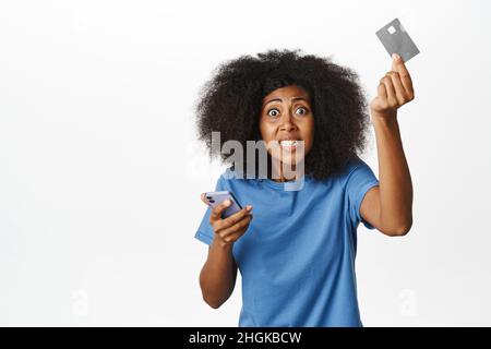 Verärgerte afrikanische Frau, die ihre Kreditkarte schrie und schüttelte, ihr Smartphone hielt, ärgerte und frustrierte, sich bei der Bank beschwerte, über Weiß stand Stockfoto