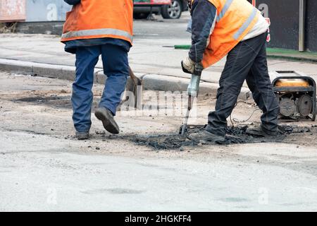 Straßenarbeiter in orangefarbenen Westen brechen auf einem reparaturbedürftigen Straßenabschnitt mittags mit einem elektrischen Presslufthammer alten Asphalt auf. Speicherplatz kopieren. Stockfoto