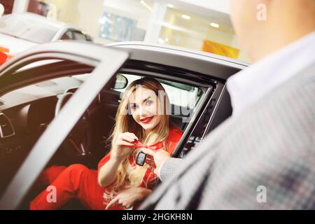 Die Hand eines Mannes in Nahaufnahme gibt die Schlüssel zu einem neuen Auto einer jungen attraktiven blonden Frau Stockfoto