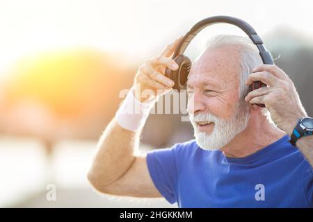 Senior lächelnder Mann mit Bart in Sportkleidung setzt Kopfhörer auf und bereitet sich auf die Bewegung in der Natur vor Stockfoto