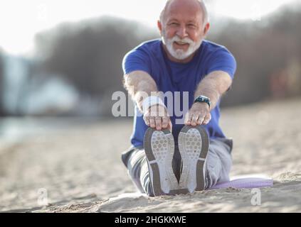 Älterer Mann mit Bart in Sportswear, der sich am Strand dehnt, während er im Sand sitzt und ein Armband und eine intelligente Uhr mit selektivem Fokus trägt Stockfoto
