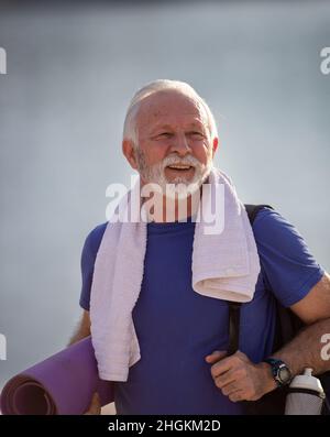 Lächelnder älterer Mann nach dem Training, der ein Handtuch um den Hals trug, während er die Yogamatte in seinem Arm und den Rucksack auf seiner Schulter hielt Stockfoto