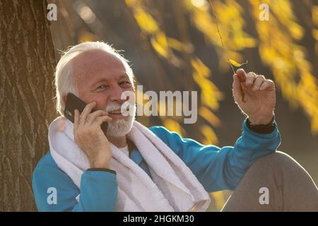 Der ältere Mann im Trainingsanzug sitzt in der Natur und redet am Telefon und entspannt sich nach dem Training, während er ein Handtuch um den Hals trägt Stockfoto