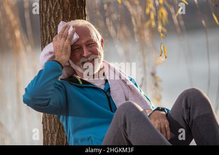 Der ältere Mann im Trainingsanzug sitzt in der Natur und lehnt sich an einen Baum und wischt sich nach dem Training mit einem Handtuch den Schweiß von der Stirn Stockfoto