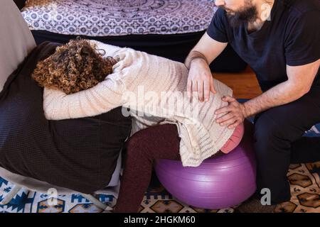 Im Chiropraktiker-Büro ist ein männlicher Praktizierender mit den Händen auf dem unteren Rücken einer Frau, die auf einem Gleichgewichtsball sitzt, zu sehen. Stockfoto
