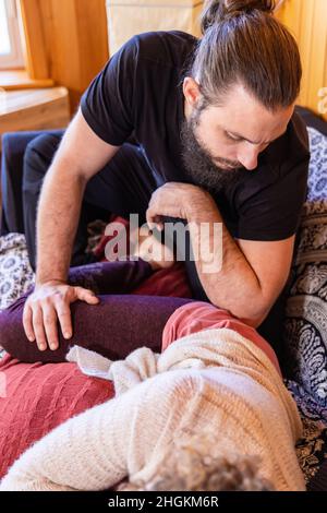 Ein kaukasischer Mann mit Bart und Haaren, die in einem Brötchen gebunden sind, führt eine Shiatsu-Massagetechnik an einer entspannten Frau durch, die im Vordergrund im weichen Fokus gesehen wird. Stockfoto