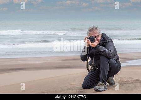 Grauhaariger erwachsener Mann mit einer Brille, die auf einer Düne kniet und ein Foto abnimmt. Außerhalb des Fokus auf Strand und Meer Hintergrund. Stockfoto