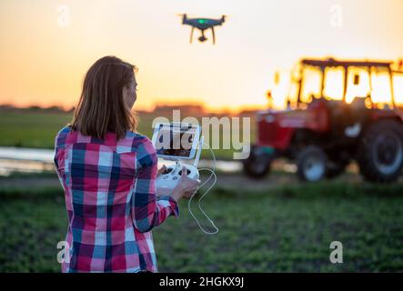 Weibliche Agronomin fliegende Drohne mit Controller-Innovation in der Landwirtschaft. Junger Bauer steht auf dem Feld mit Traktor im Hintergrund Stockfoto