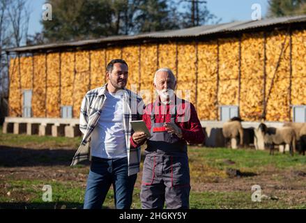 Glückliche Farmarbeiter, die auf einem Bauernhof mit einem digitalen Tablet zusammenarbeiten. Ein älterer Mann zeigt etwas in der Ferne. Stockfoto
