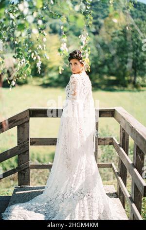 Braut in einem langen weißen Korbkleid steht auf der Terrasse in der Nähe des Geländers Stockfoto