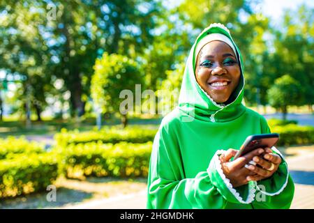 lateinamerikanische hispanic arabische Frau in grünem muslimischen Kleid mit modernem hellen Make-up und Nasenpiercing Blick auf Telefonbildschirm im Freien Stockfoto